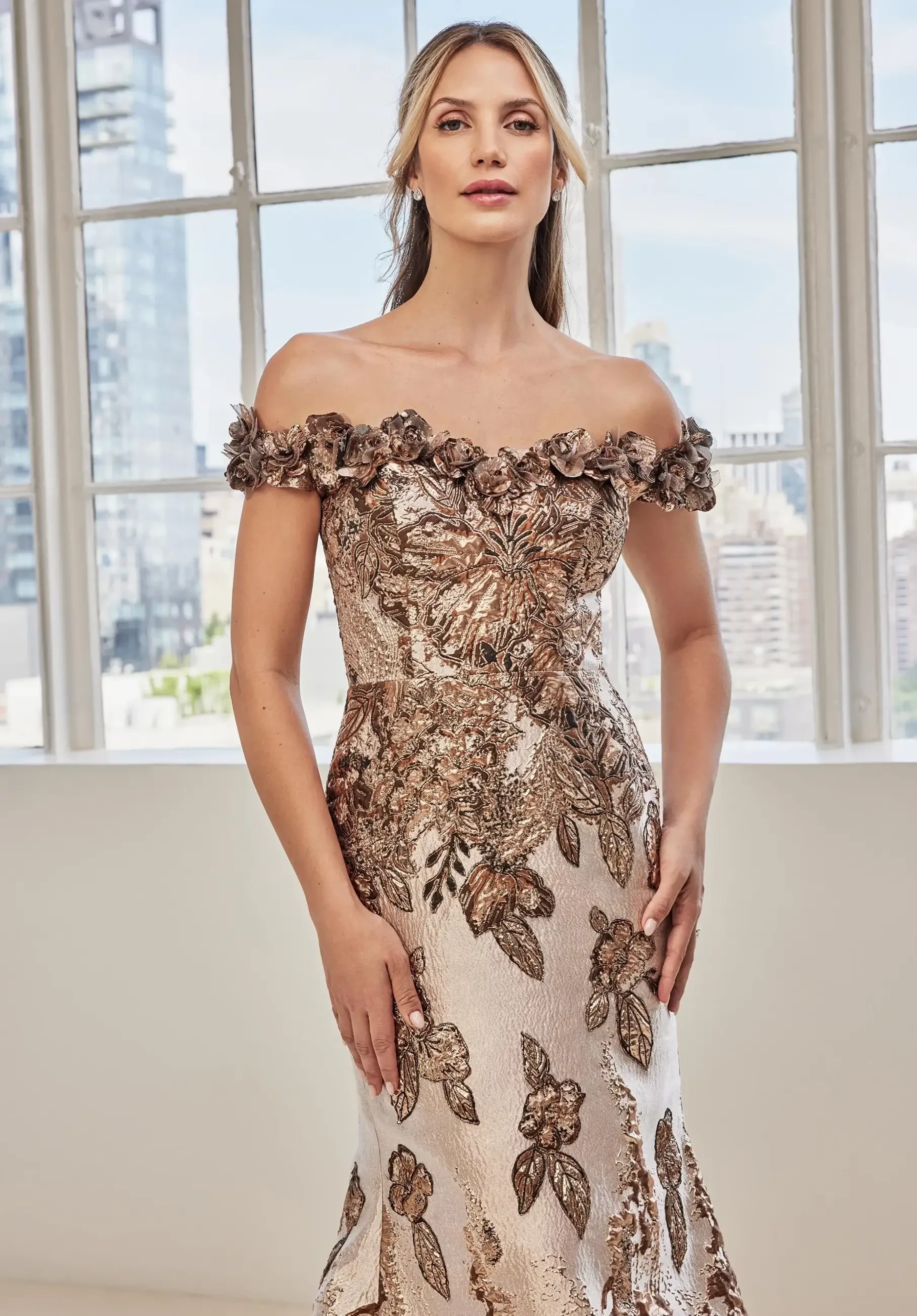 A woman stands in front of large windows with a cityscape backdrop. She wears an elegant off-the-shoulder gown with floral embroidery, exuding grace and confidence.