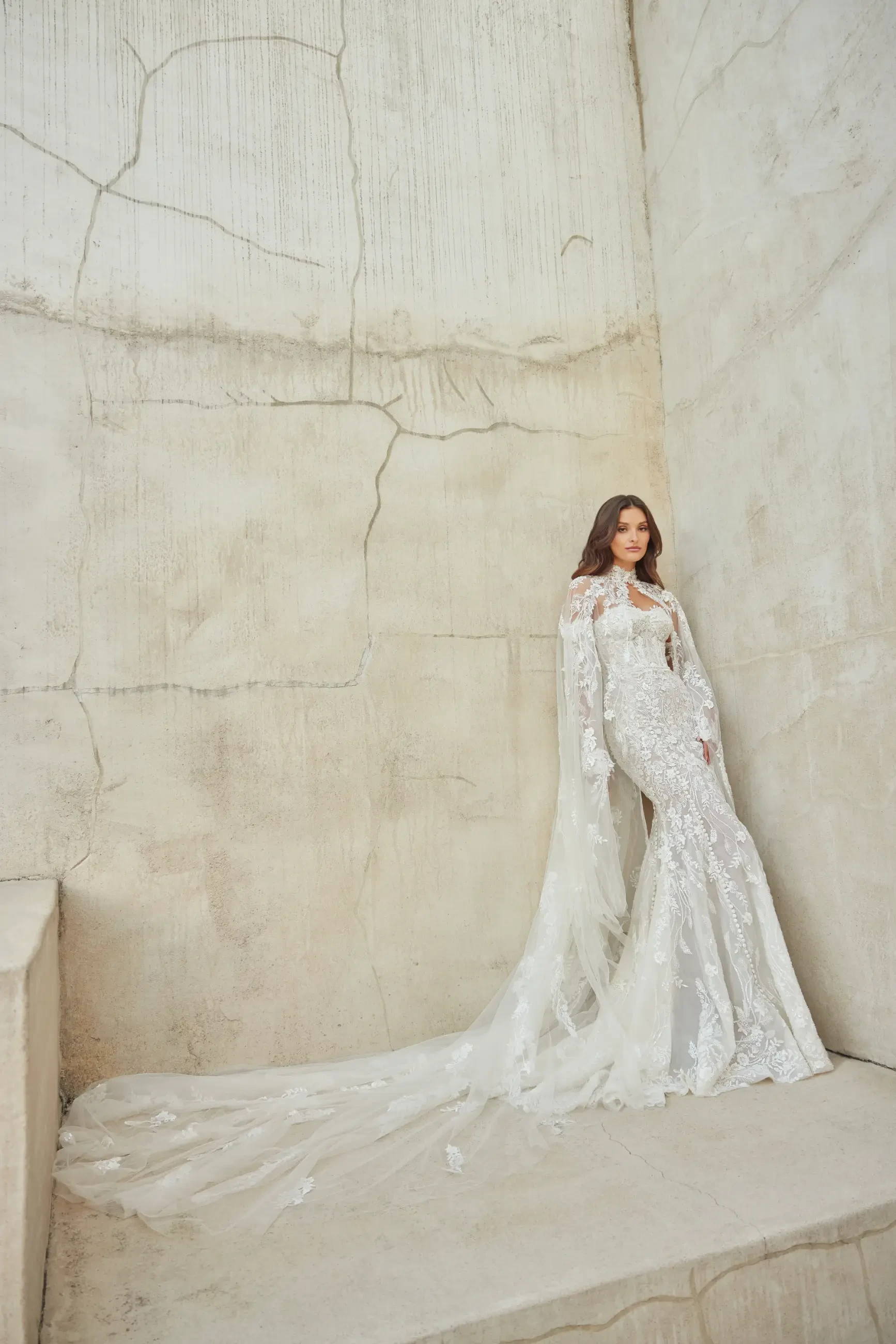 Model is wearing the Lotus gown, a stone-colored strapless mermaid dress with hand-sewn lace appliques, beads, sequins, a sweetheart neckline, and a detachable beaded tulle cape.