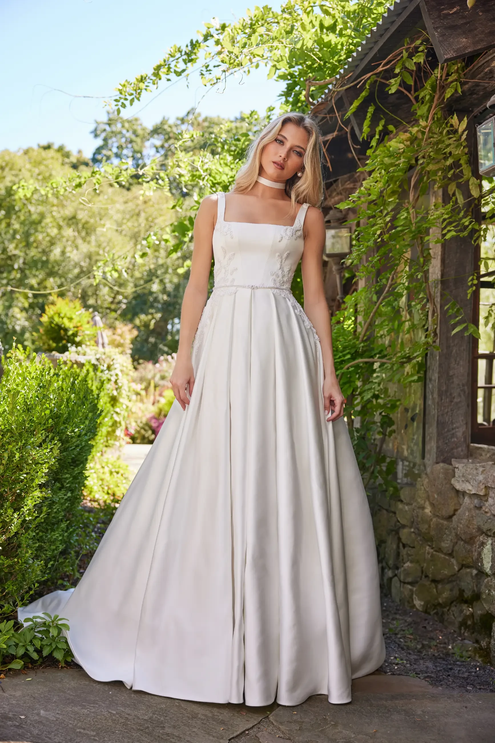 Model is wearing the Daphne gown, an ivory satin ballgown with a square neckline, sleeveless design, pearl accents at the waist and sides, a lace-up back, and a satin choker.