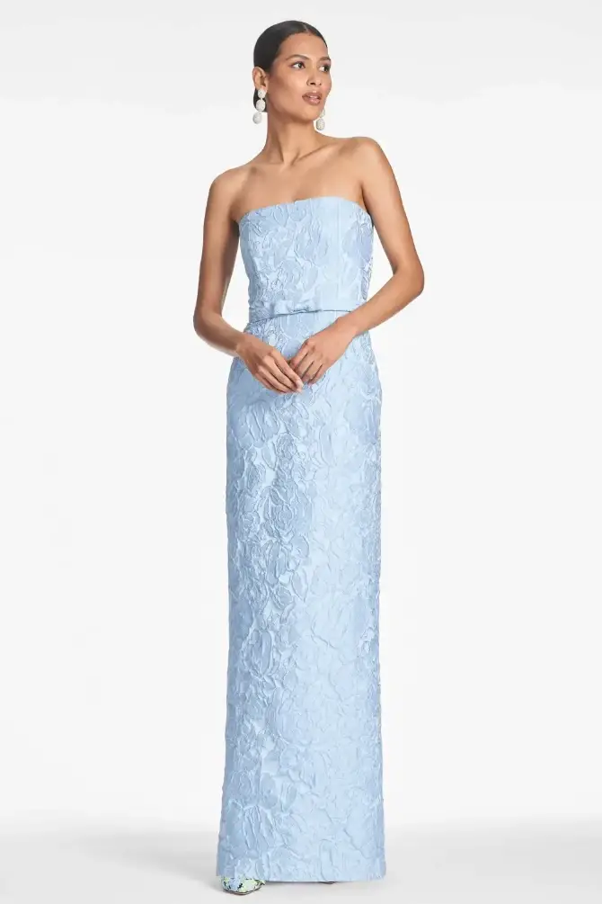 Woman in elegant strapless blue gown with textured floral pattern, standing confidently. She wears pearl earrings, exuding sophistication and grace.