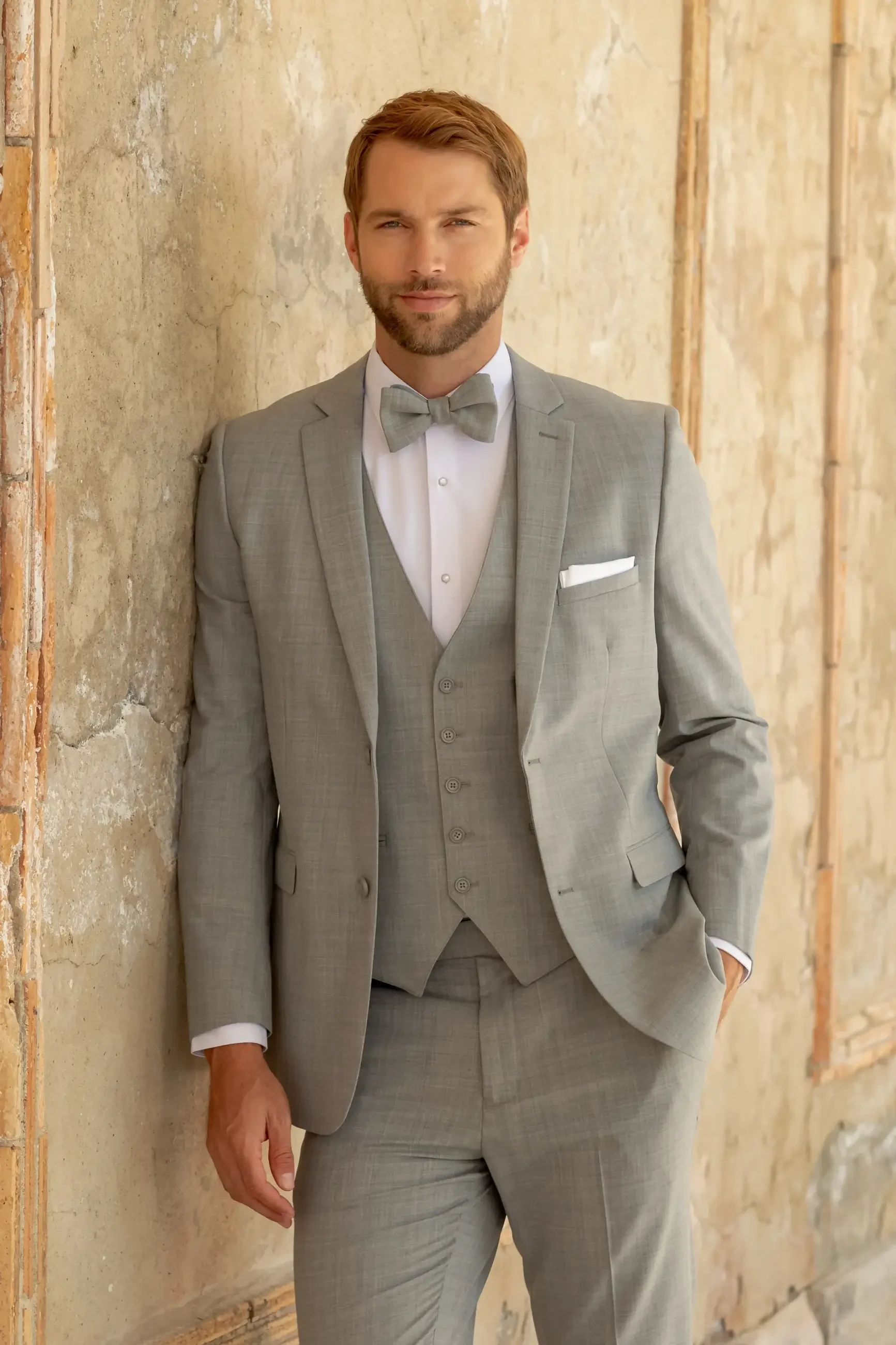 A man in a light gray suit with a bow tie stands against a textured wall, looking confidently at the camera.