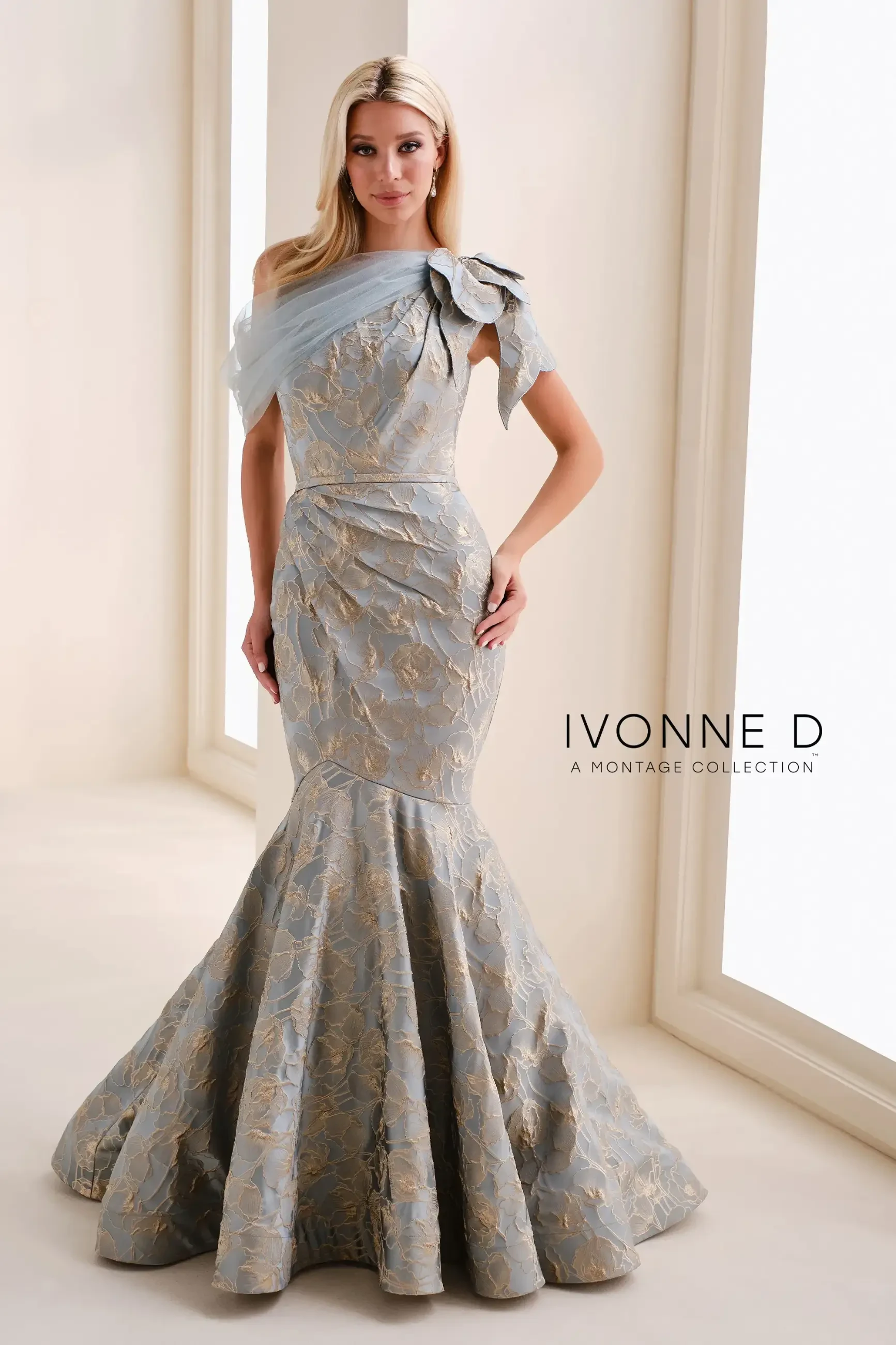A woman is wearing a formal gown with an elegant design, featuring a shoulder detail and a flared bottom. The dress has intricate patterns and is made of light fabric, set against a softly lit background. The brand name 
