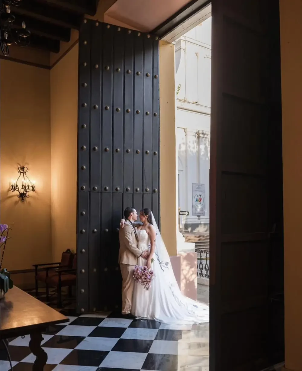 A bride and groom share a romantic moment inside a building, framed by an open door with large black panels, while sunlight streams in. The floor features a checkered pattern.