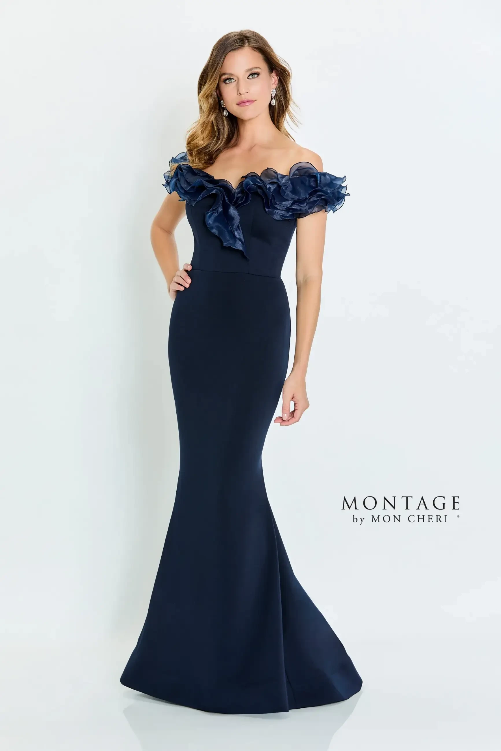 A woman in a navy off-the-shoulder evening gown with ruffled details stands confidently against a white background, exuding elegance and sophistication.