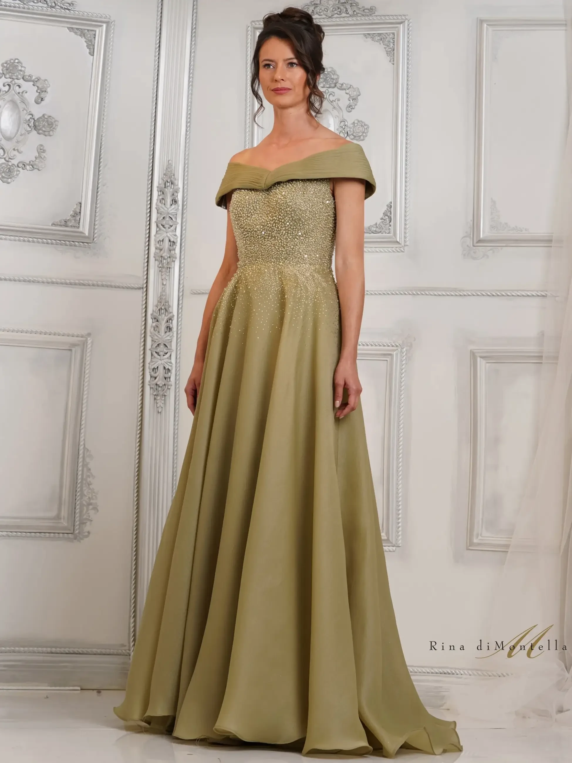 A woman wearing an off-the-shoulder, olive-green evening gown with embellishments, standing in front of a decorative wall.