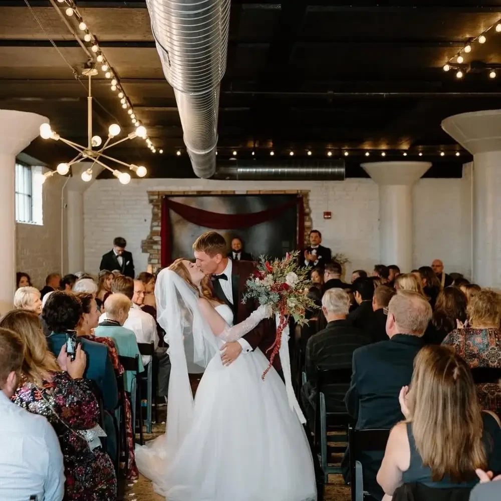 A wedding ceremony taking place indoors, featuring a couple exchanging vows, surrounded by seated guests in formal attire. The venue has a modern aesthetic with exposed beams and decorative lighting.