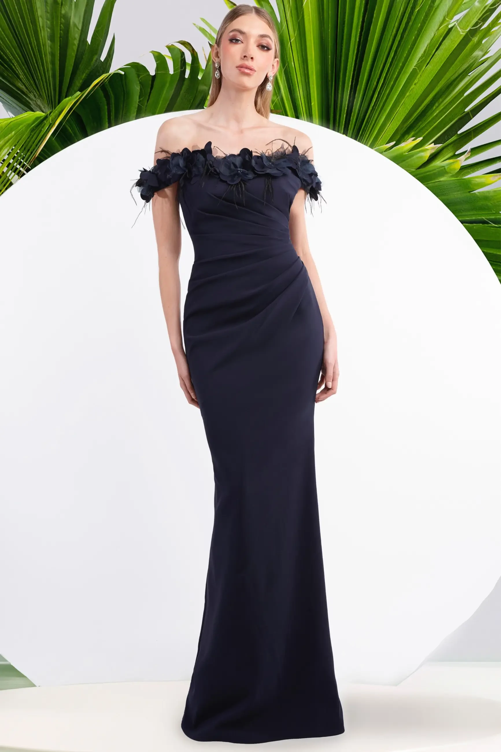 A woman in an elegant, off-the-shoulder navy gown with floral details stands confidently. Green palm leaves form a lush backdrop, creating a stylish, serene ambiance.